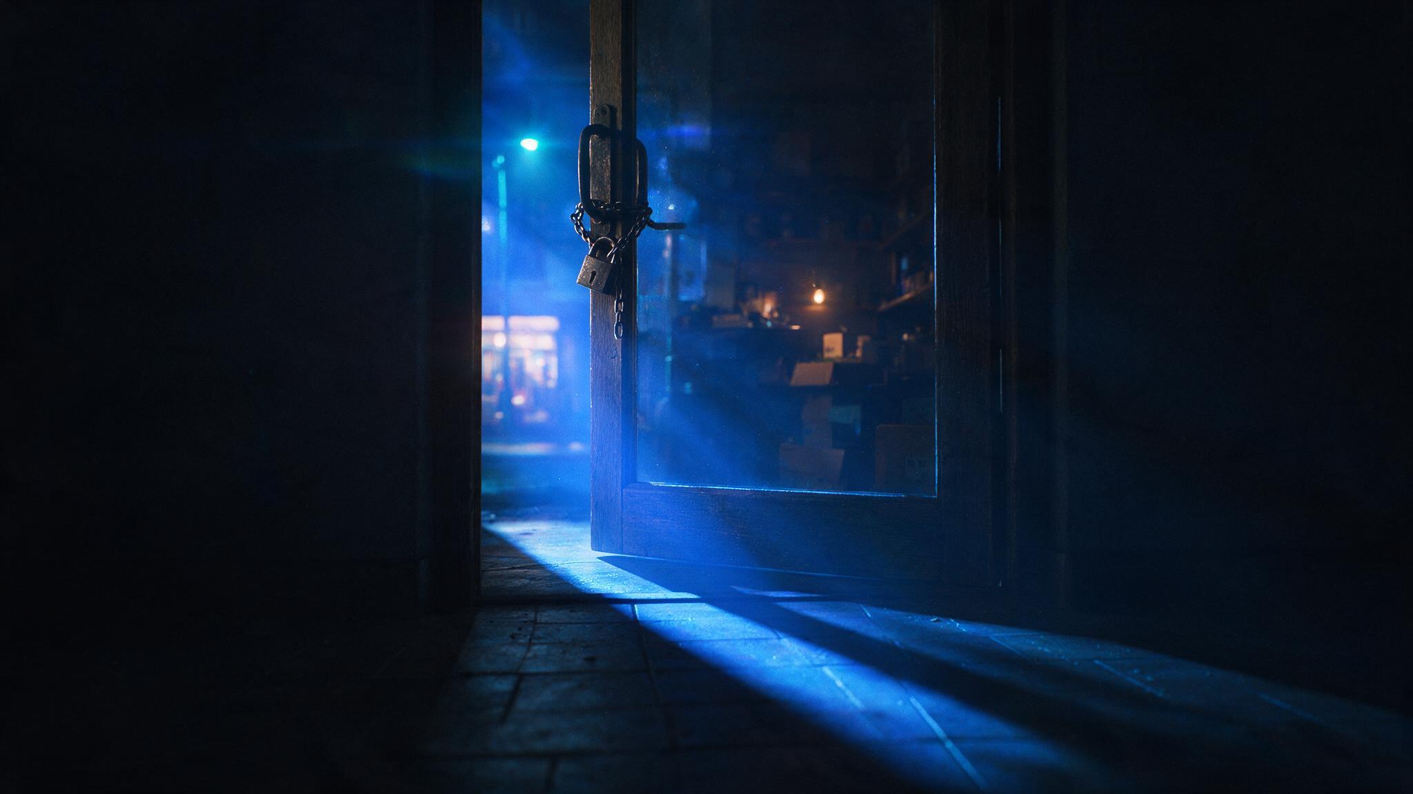A shop's front door seen from inside at night, a padlock and chain on the inside handle, the key missing